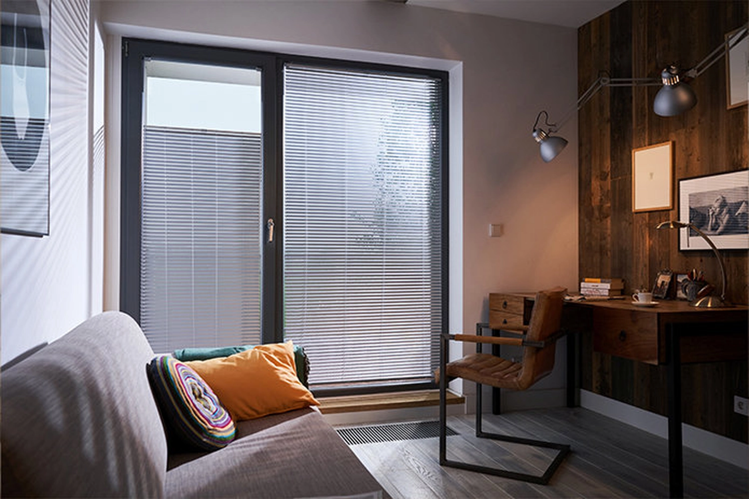 Cybulski Trockenbau - Bau und Fensterbauer - Ein gemütliches, modernes Zimmer mit einem grauen Sofa mit orangefarbenem Kissen, einem Schreibtisch mit braunem Ledersessel und großen Fenstern mit Jalousien.