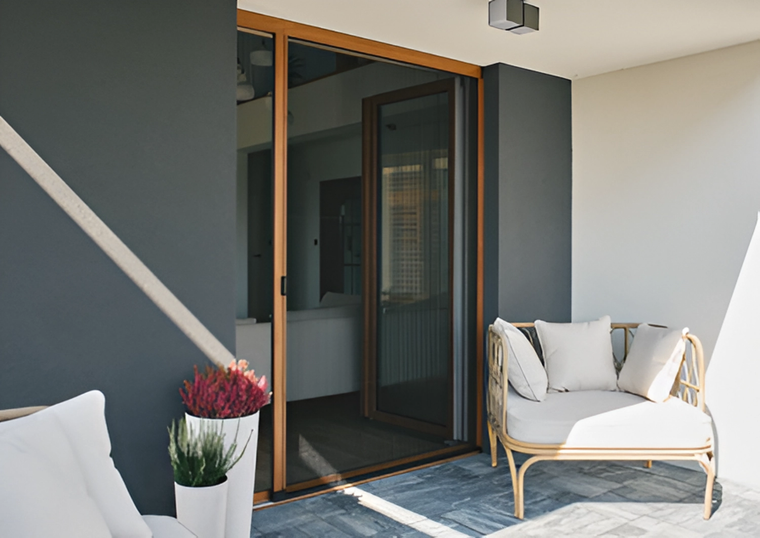 Cybulski Trockenbau - Bau und Fensterbauer - Eine moderne Terrasse mit einem Rattan-Gartensofa mit weißen Kissen, neben großen Glasschiebetüren mit Holzrahmen.